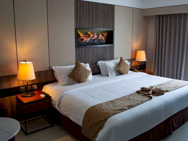 A cozy hotel room features a large bed with white linens, decorative pillows, and warm lighting from bedside lamps, creating a welcoming ambiance.