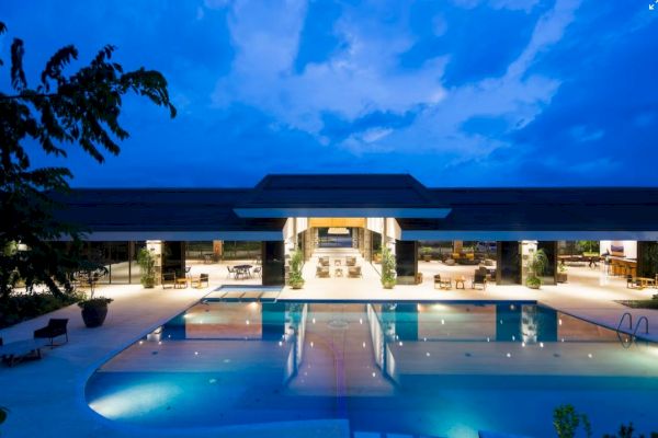 The image shows a modern building with an illuminated pool in the foreground under a twilight sky.