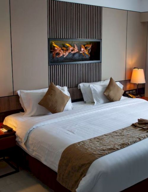 The image shows a hotel room with a large bed, decorative pillows, two bedside lamps, and a framed picture on the wall.