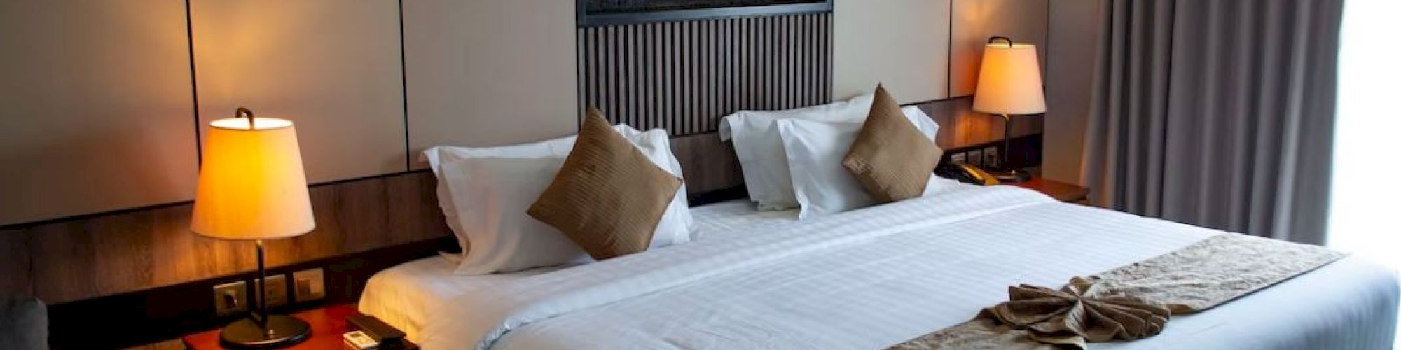 The image shows a neatly arranged hotel room with a large bed, pillows, lamps on side tables, and a decorative piece on the wall above the bed.