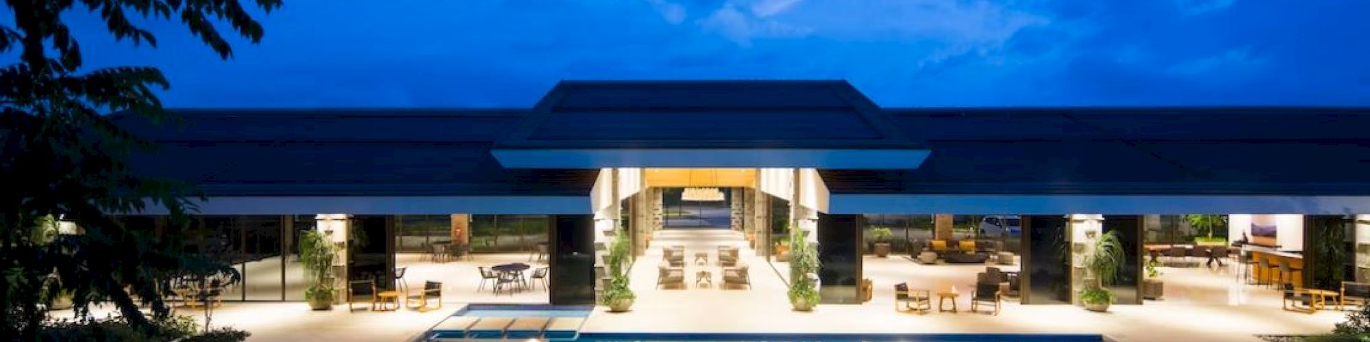 A luxurious modern building with a large illuminated pool at night, surrounded by elegant seating areas and set against a blue sky.