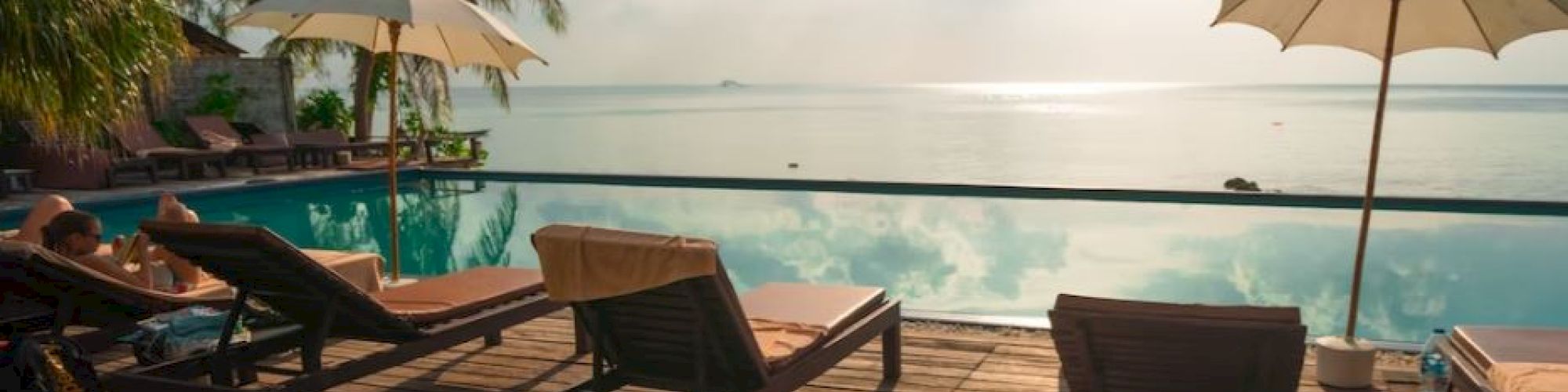 A sunny deck by a pool with lounge chairs and umbrellas, overlooking calm waters and a clear sky with scattered clouds.