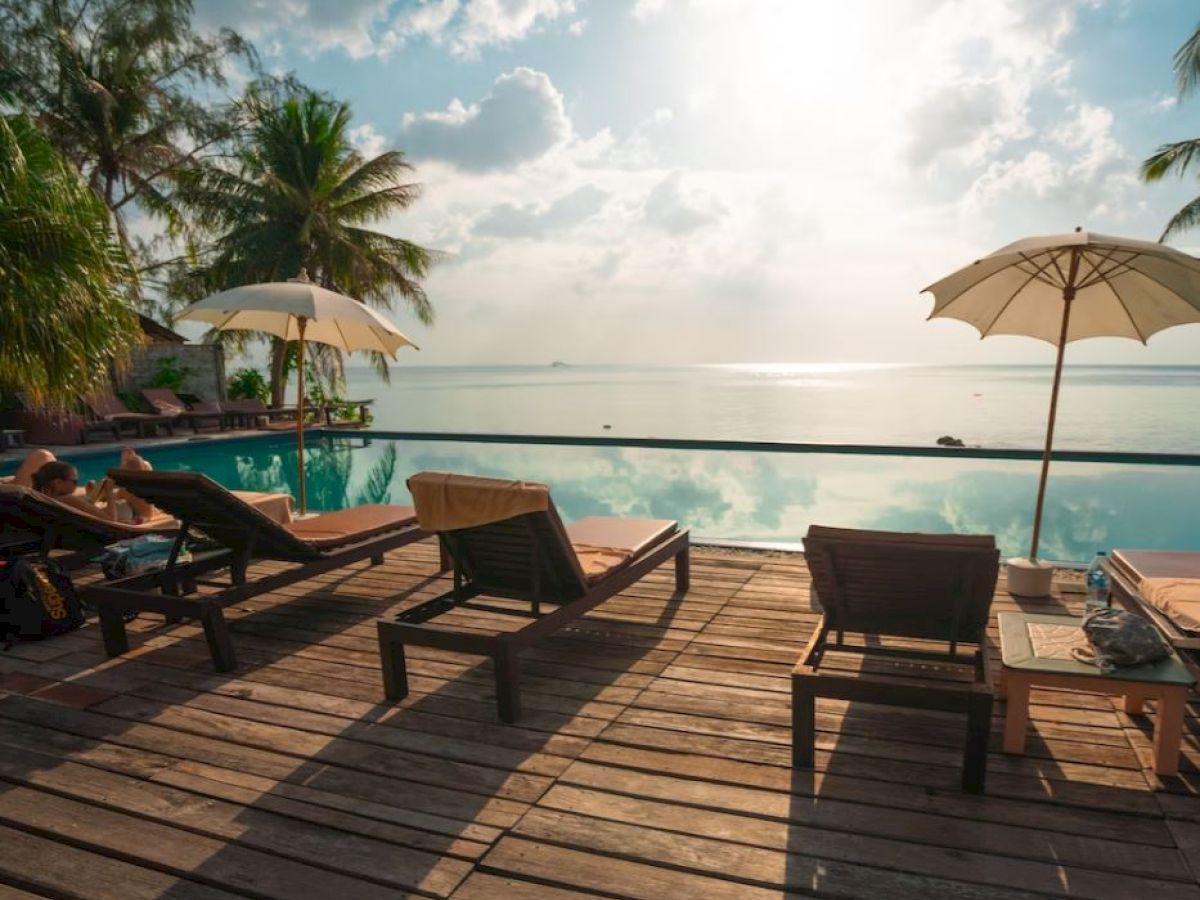 Poolside deck with lounge chairs and umbrellas overlooking a calm, scenic waterfront under a partly cloudy sky with palm trees nearby.