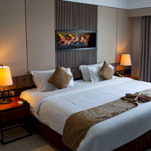 A neatly arranged hotel room features a large bed with white linens, brown accent pillows, bedside lamps, and a decorative wall panel.
