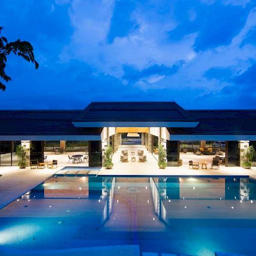 The image shows a modern building with a lit swimming pool at night, under a cloudy sky, creating a serene atmosphere.