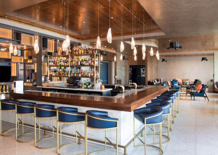 A modern bar with a wooden countertop, blue barstools, stylish lighting, and shelves filled with bottles. Cozy seating is in the background.