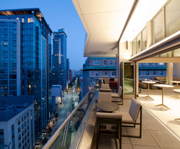 A modern rooftop terrace with seating, overlooking an urban cityscape of high-rise buildings during the evening.