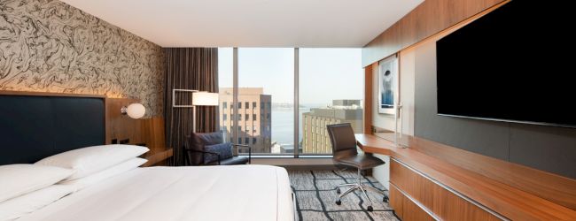 A modern hotel room with a large bed, wooden furniture, flat-screen TV, and a desk overlooking a city view through a large window.