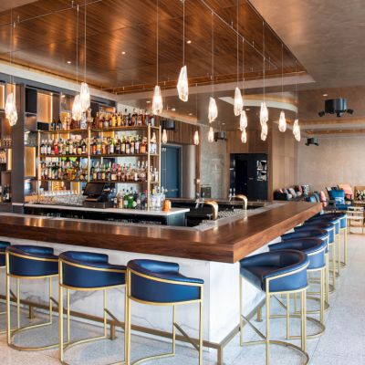 The image shows a modern bar with a row of blue stools, hanging lights, and a well-stocked back bar. Seating area visible in the background.