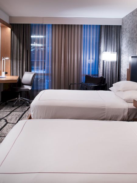 The image shows a hotel room with two beds, a wall-mounted TV, desk, chair, and lamps, featuring a modern design and decor.