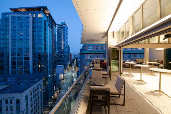 A modern rooftop terrace at dusk with tables and chairs overlooking a cityscape of tall glass buildings under a deep blue sky.