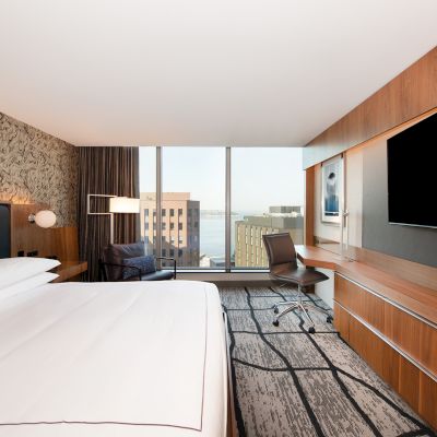 A modern hotel room with a large bed, wall-mounted TV, desk, chair, and window offering a city view in the background.