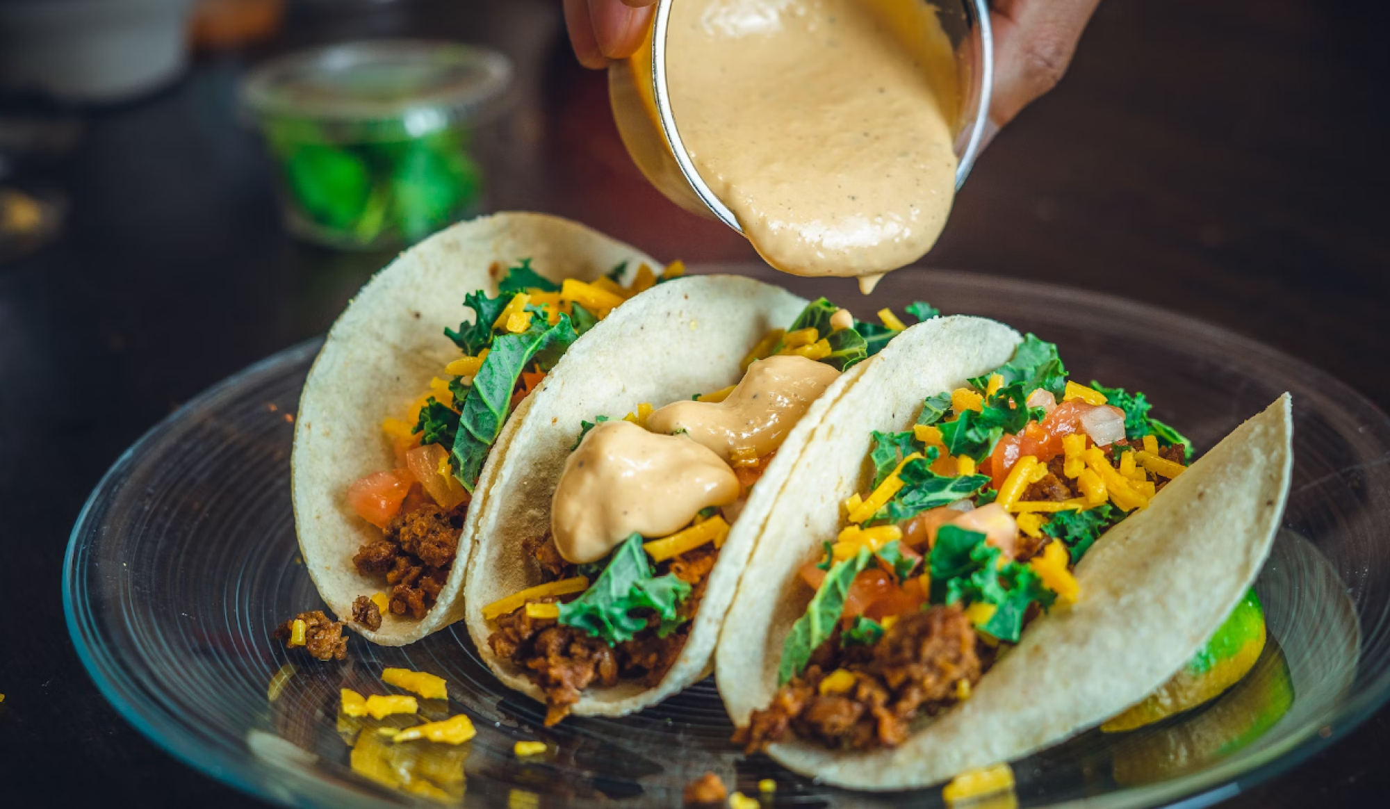 Three tacos filled with ground meat, cheese, lettuce, and tomatoes are on a plate. Sauce is being poured over them.