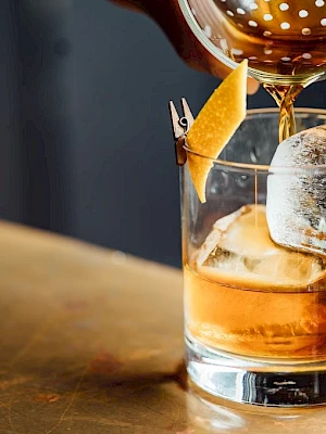 A drink being poured into a glass with ice and a twist of citrus peel, possibly preparing a cocktail.