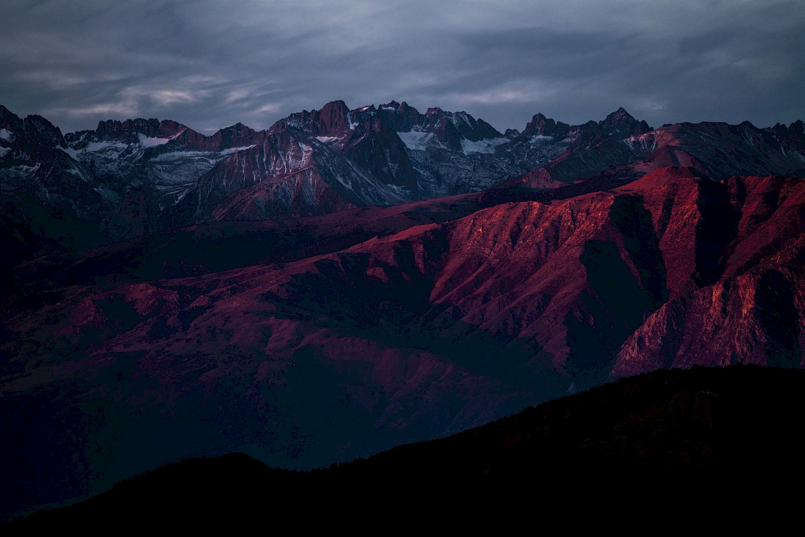 A mountain range with reddish hues under a cloudy sky, likely at sunrise or sunset.
