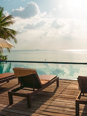 A serene beachside setting with lounge chairs, umbrellas, and a pool overlooking the ocean, surrounded by palm trees under a sunny sky.
