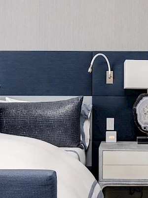 A modern bedroom features a bed with blue pillows, a white nightstand, a lamp, and a reading light attached to the headboard.