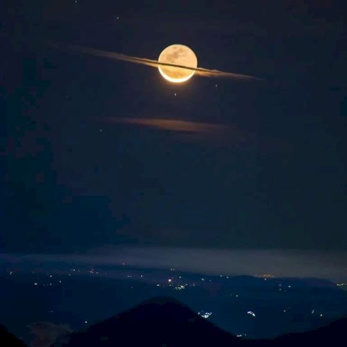 A glowing full moon with clouds resembling Saturn's rings, above a dark mountainous landscape with distant city lights.