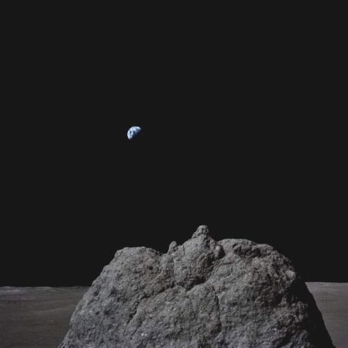 Earth is seen rising above the lunar surface, with a rocky foreground on the Moon, against the blackness of space.