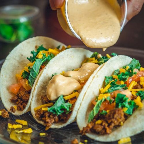 Three tacos filled with ground meat, cheese, lettuce, and tomatoes, being topped with a creamy sauce on a plate.