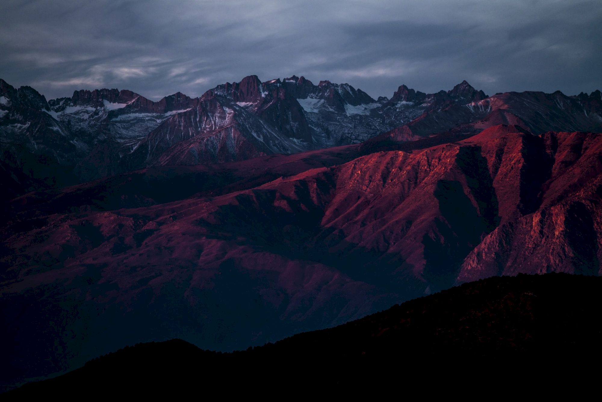 Mountain range with snow-capped peaks under a cloudy sky, illuminated by red sunlight.