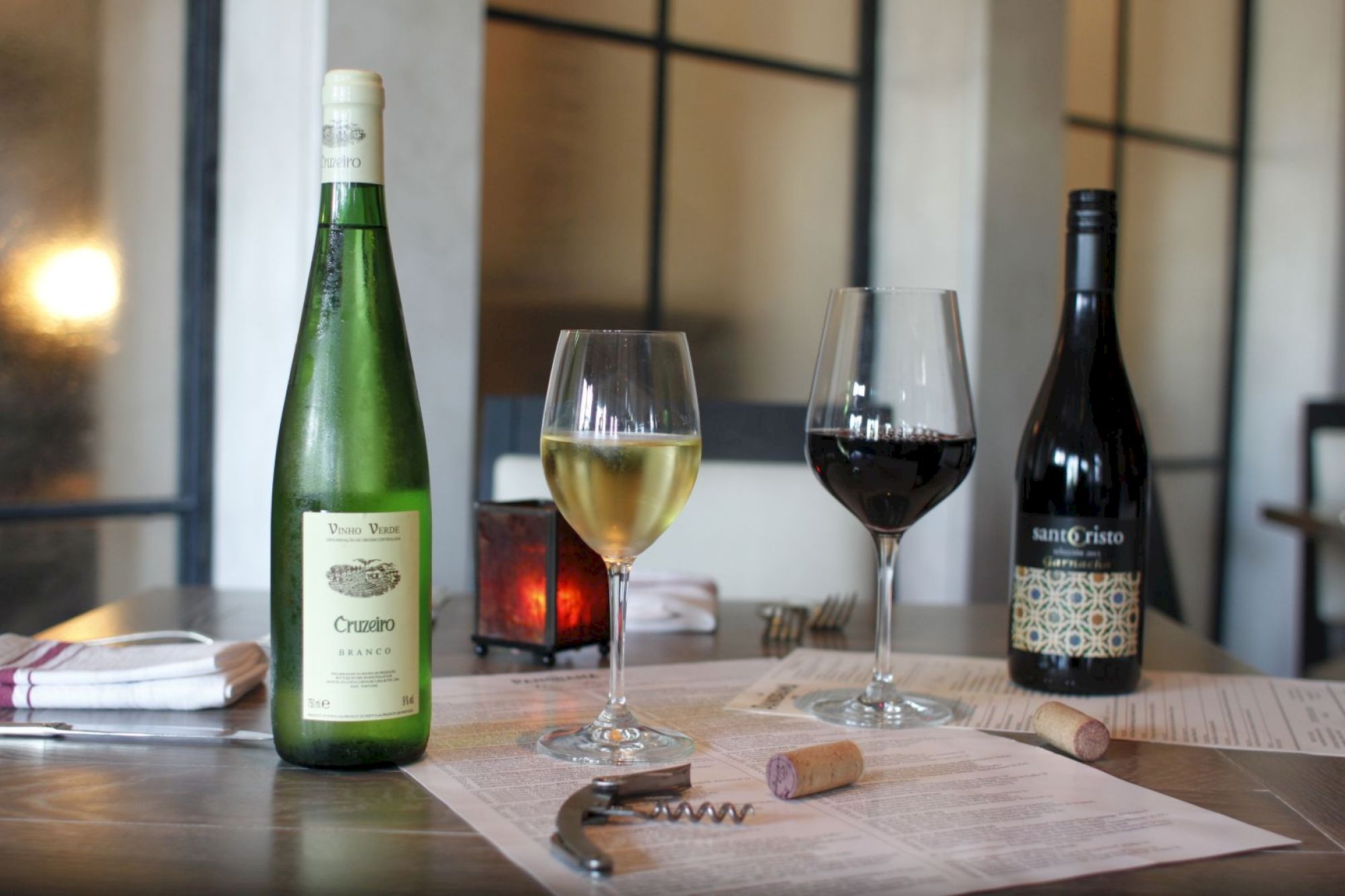 Two wine bottles, a glass of white and red wine, a corkscrew, corks, and menus are on a table in a dimly lit setting.