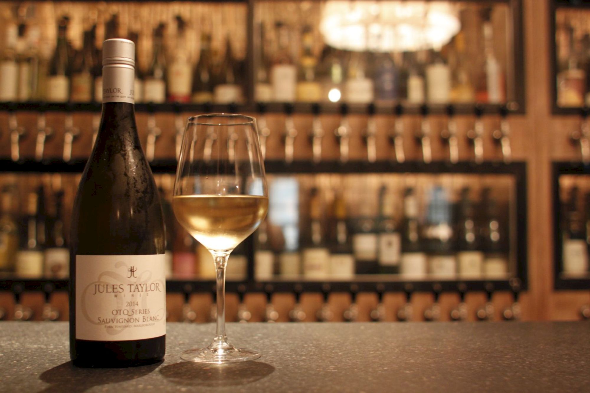 A bottle of Jules Taylor wine and a filled wine glass on a counter, with shelves of various wines in the blurred background.