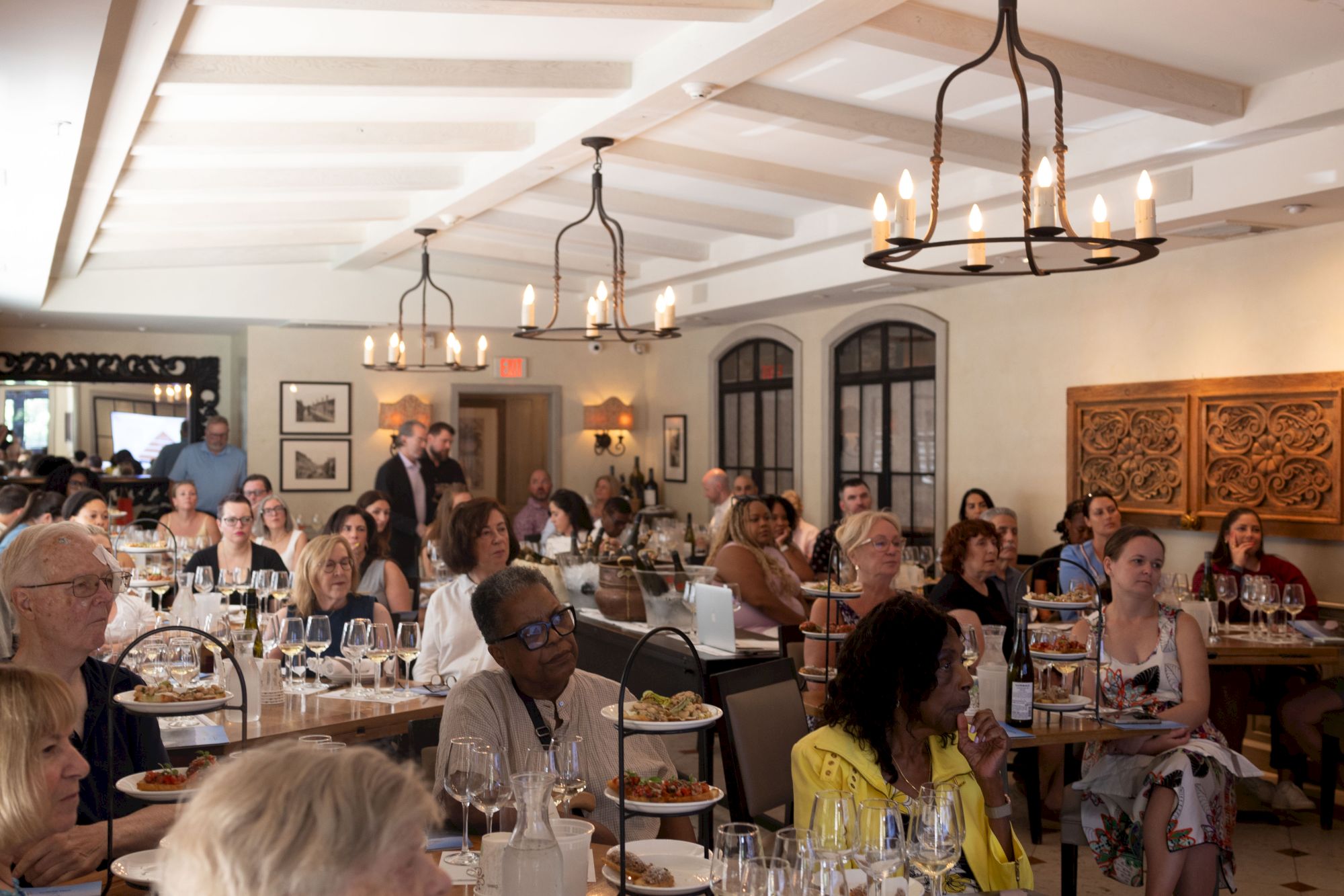 Inside a banquet hall, diners sit at tables under hanging candle chandeliers, chatting during a formal meal.