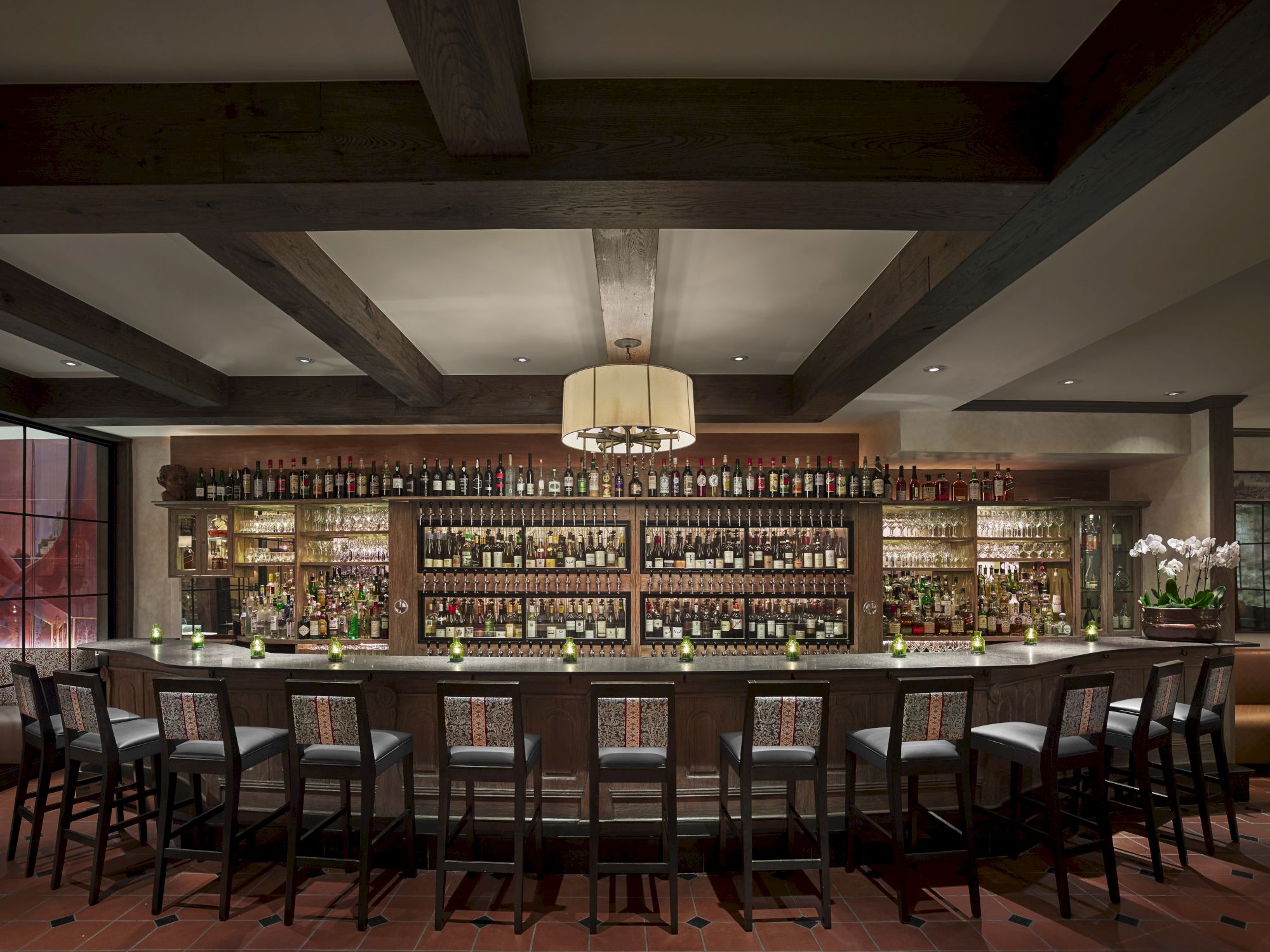 The image shows a stylish bar with a long counter, numerous bottles displayed on shelves, and several bar stools arranged in front of it.