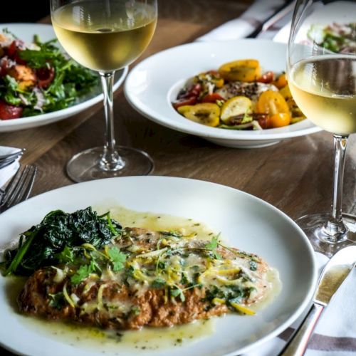 The image shows a dining setup with plates of gourmet food, including a saucy dish with greens, salads, and glasses of white wine on a table.
