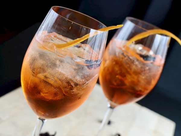 Two glasses filled with a refreshing orange-hued cocktail, topped with ice and garnished with a long citrus peel.