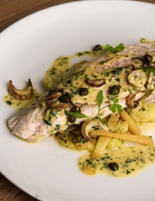 A plated fish dish with a creamy herb sauce, garnished with capers, mushrooms, and herbs on a white plate.