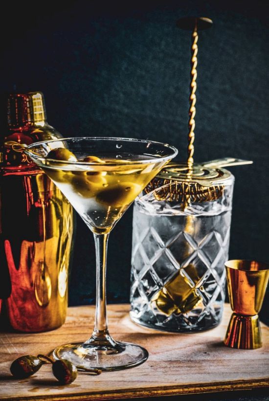 A martini with olives, a crystal glass with a stirrer, a cocktail shaker, jigger, and olives on a wooden surface are displayed.
