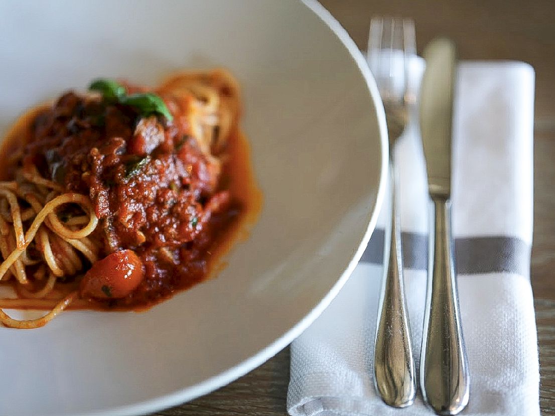 A bowl of spaghetti with tomato sauce and meat, placed beside a knife and fork on a napkin.