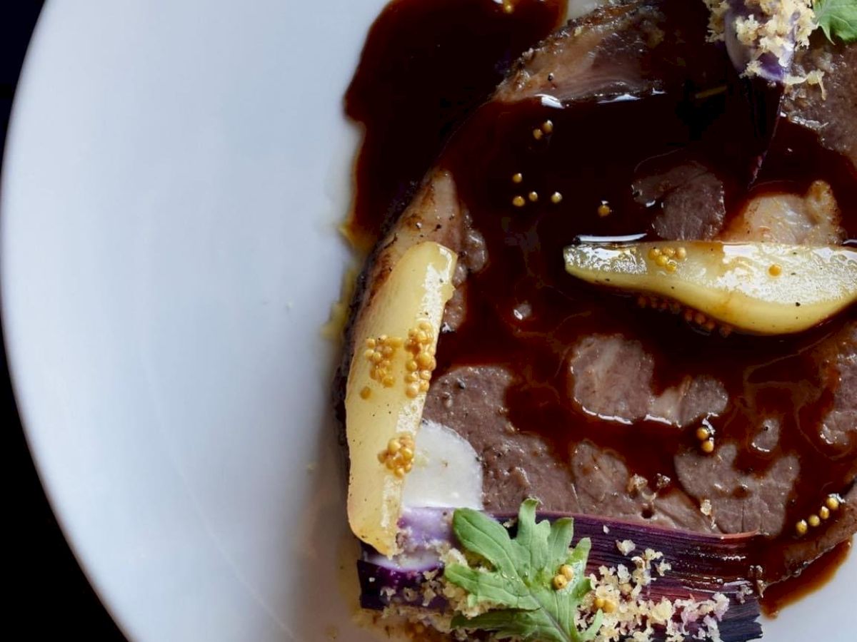 A gourmet dish featuring sliced meat with sauce, garnished with vegetables, herbs, and crumbs, artistically arranged on a white plate.