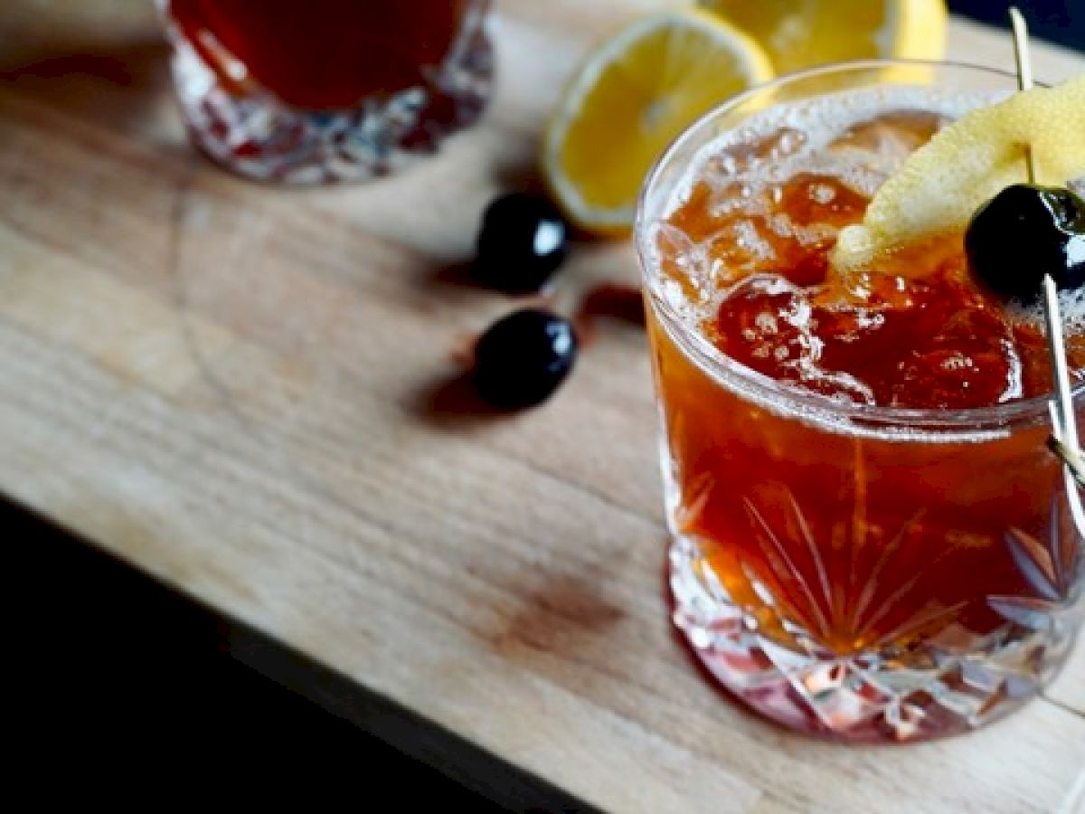 A cocktail garnished with a lemon twist and cherry on a wooden board, alongside another drink, cherries, and lemon slices.