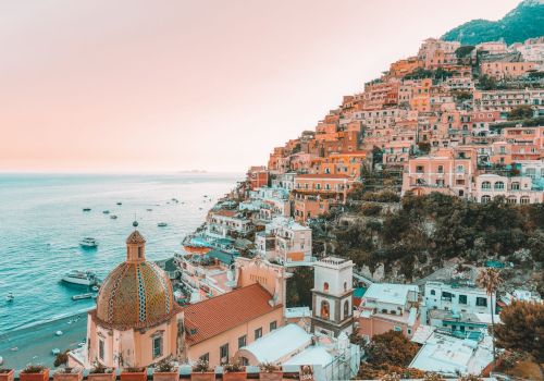 A coastal hillside town with colorful buildings overlooking the sea, boats in the water, and a dome-topped church in view.