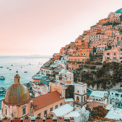 A coastal hillside town with colorful buildings overlooking the sea, boats in the water, and a dome-topped church in view.
