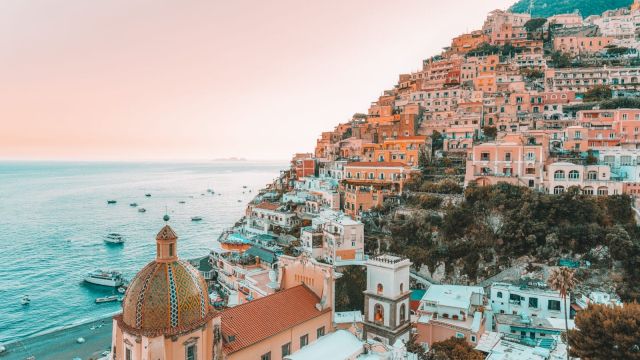 A coastal hillside town with colorful buildings overlooking the sea, boats in the water, and a dome-topped church in view.