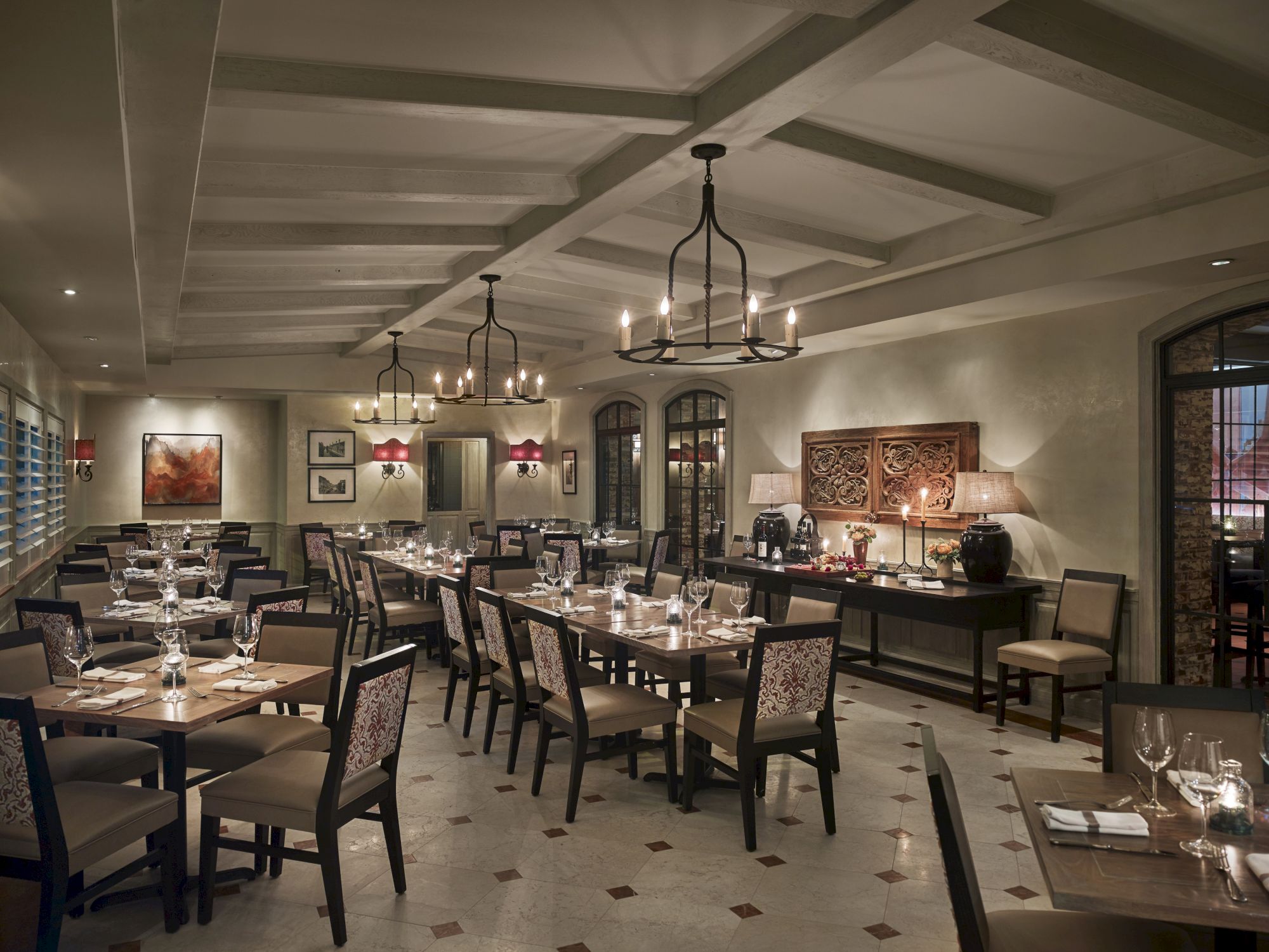 The image shows an elegant, empty restaurant with neatly set tables, chandeliers, and framed artwork on the walls.