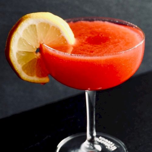 A red cocktail in a coupe glass, garnished with a slice of lemon on the rim, set against a dark background.