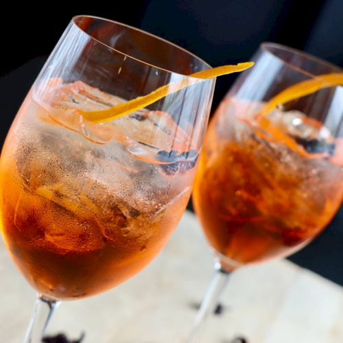 Two glasses of orange-tinted cocktails with ice and orange twists as garnish, placed on a light-colored surface.