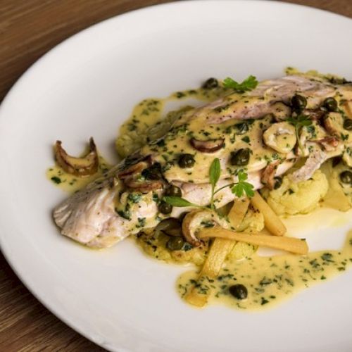 Grilled fish with a creamy herb sauce, topped with capers and mushrooms, served on a white plate.