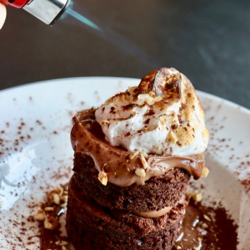 A dessert with chocolate layers and cream is being torched on top, garnished with nuts and cocoa powder, on a white plate.