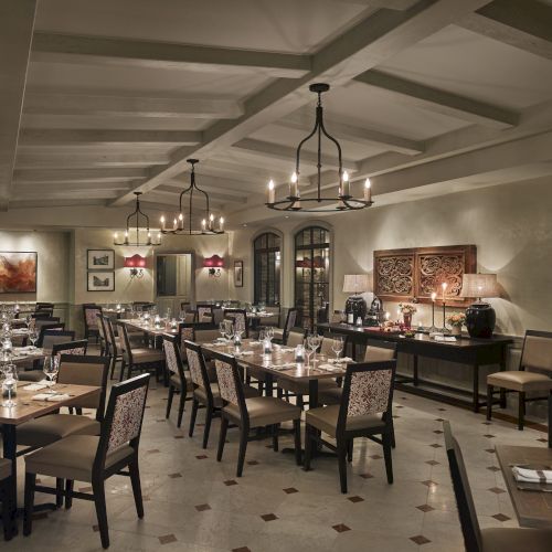 Luxurious dining room with elegant chandeliers, wooden tables, and chairs set for guests, featuring art on the walls and warm lighting.