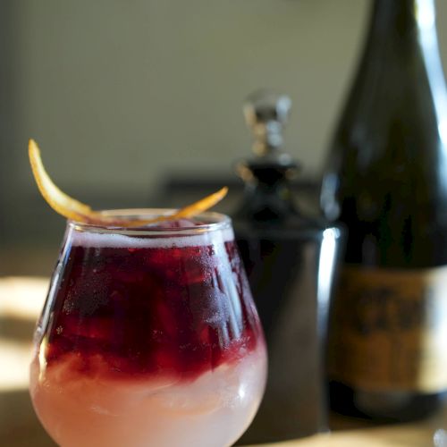 A cocktail in a glass with layered colors and a citrus twist, accompanied by bottles in the background on a wooden table.