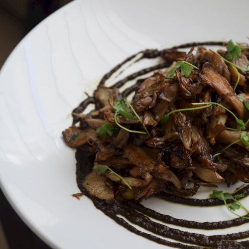 A white plate with sautéed mushrooms, garnished with greens, on top of a decorative dark sauce swirl.