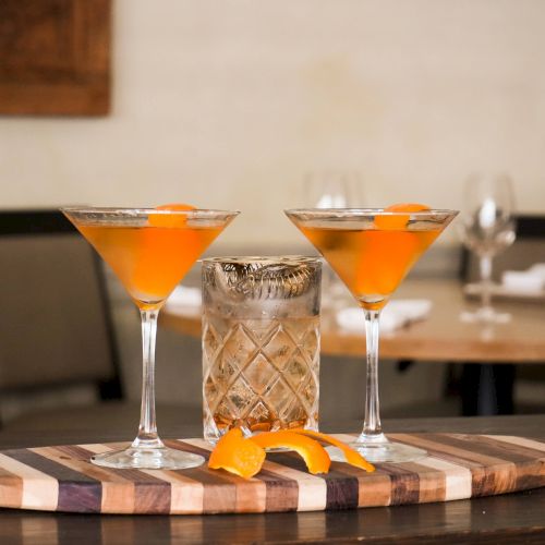 Two orange cocktails in martini glasses on a wooden board with a glass and orange peel between them, set against a restaurant backdrop.