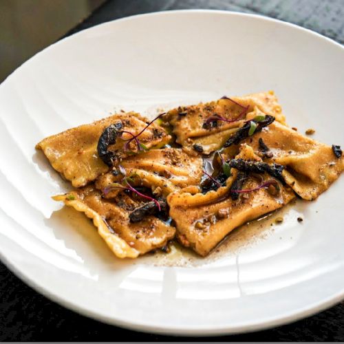 The image shows a white plate with ravioli topped with herbs and a sprinkle, likely spices or breadcrumbs.