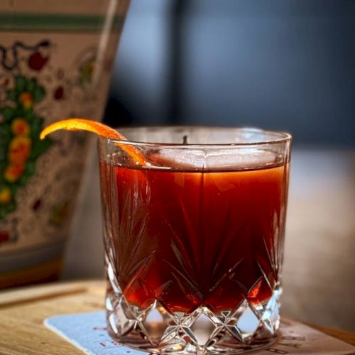 A glass with a dark red cocktail on a coaster, garnished with an orange twist, beside a decorative vase.